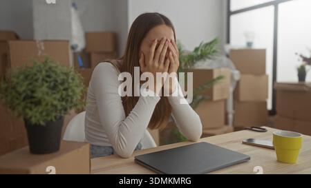 Femme avec la main sur la joue regardant l'ordinateur portable dans le bâtiment entouré de boîtes en mouvement emballées, plante, et tasse sur la table ; stress de jour en mouvement. Banque D'Images