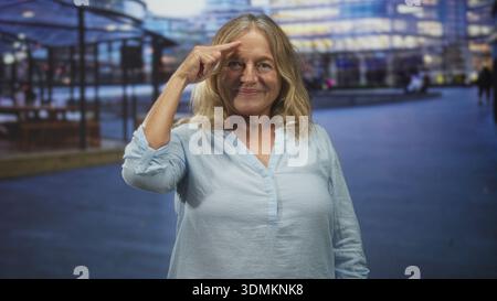 Femme blonde senior sourit et pointe l'index droit à la tempe avec la main visible tout en se tenant debout à l'extérieur de l'entrée de l'immeuble de bureaux moderne au crépuscule ; t Banque D'Images