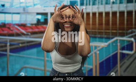 Femme brune dans un bureau blanc et un Jean, mains formant des jumelles sur les yeux tout en souriant à une terrasse de la piscine intérieure avec chaises longues et balustrades ; pl Banque D'Images