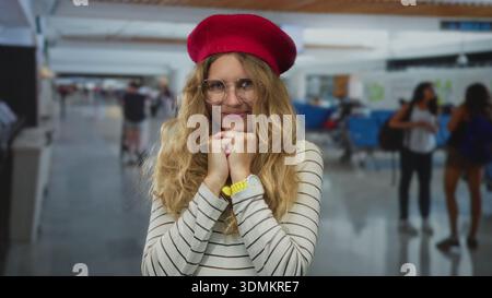 Femme se reposant les mains sous le menton et souriant à l'aéroport, jeune modèle blond ajustant les lunettes et béret rouge sur le pull rayé. Banque D'Images