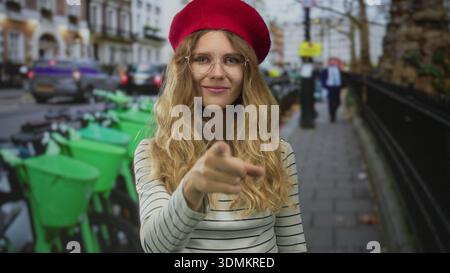 Femme utilise son doigt pour pointer la rue animée tandis qu'une jeune voyageuse blonde dans un béret rouge et des lunettes explore une scène urbaine. Banque D'Images