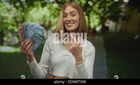 Jeune femme blonde dans un parc détenant des billets de banque hongrois en forint à l'extérieur, exprimant le bonheur et le succès financier, une verdure vibrante dans le backgrou Banque D'Images