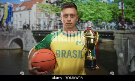 Maillot homme au brésil tient le basket-ball et le trophée, la main droite saisissant le ballon tout en se tenant debout dans la rue ; fierté de la victoire. Banque D'Images