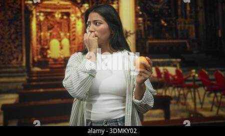 Femme tenant une orange tout en appuyant les doigts sur les lèvres dans une allée de bâtiment ornée, les yeux fermés et clouant avec chemise décontractée et jeans visible ; an Banque D'Images