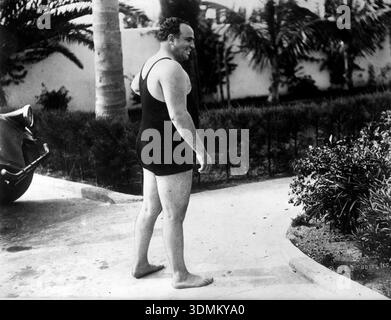 Al Capone, le tristement célèbre gangster américain, est capturé dans cette photographie monochrome vintage de 1921. Il est vu dehors dans un maillot de bain sombre, souriant et regardant par-dessus son épaule, avec un feuillage tropical et ce qui semble être une aile de voiture en arrière-plan. Banque D'Images