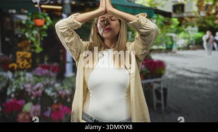 Jeune femme presse les paumes ensemble dans la prière au milieu d'un marché de fleurs de rue occupé tenant les yeux fermés et la posture calme ; sérénité pleine conscience. Banque D'Images