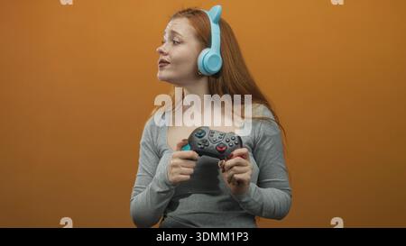 Jeune femme tenant la manette de jeu et pointant le doigt tout en portant des écouteurs d'oreille de chat bleu en studio ; ludique. Banque D'Images