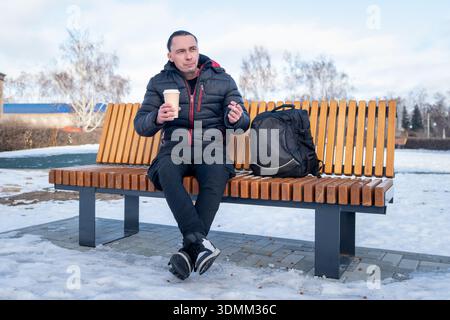 homme assis sur un banc en hiver, buvant du café et fumant une cigarette. Banque D'Images