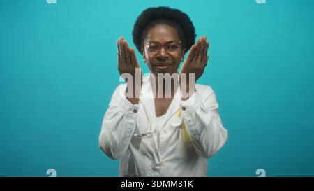 Femme médecin en blouse de laboratoire blanche et stéthoscope, portant des lunettes, des tasses mains sur le visage puis ouvre les paumes sur fond de studio turquoise ; calme resur Banque D'Images
