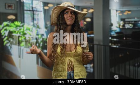 Jeune femme hispanique dans le chapeau de soleil pointe doigt et tient popsicle saupoudré à l'intérieur du bâtiment portant un haut crochet jaune et jeans ; l'été insouciant. Banque D'Images