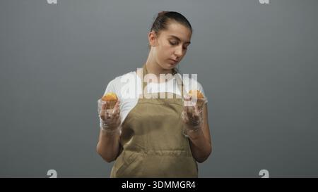 Femme tenant deux muffins avec les mains gantées et portant un tablier dans le studio, regardant vers le bas les pâtisseries ; contemplation. Banque D'Images