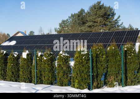 Panneau solaire couvrant un toit résidentiel derrière une haie à feuilles persistantes et une clôture en métal vert, neige profonde sur le sol, concept d'énergie propre dans la banlieue hivernale Banque D'Images