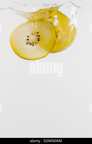 Deux tranches de kiwi jaunes tombant dans l'eau, isolées sur blanc. Banque D'Images