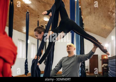 Instructeur masculin assistant un enfant dans la pratique aérienne de yoga sur des sangles d'hamac dans un studio de fitness. Banque D'Images