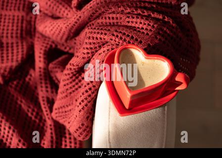 Tasse de café en forme de coeur sur fauteuil avec couverture rouge. Nature morte confortable pour la Saint-Valentin Banque D'Images