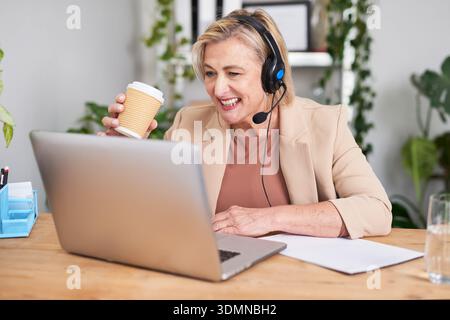 Professionnel souriant avec micro-casque/oreillette travaillant sur ordinateur portable et tasse à café au bureau Banque D'Images