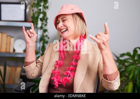 Femme senior joyeuse célébrant au travail avec Pink Hat et Floral Lei au bureau Banque D'Images