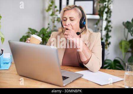 Femme d'affaires avec casque travaille au bureau d'ordinateur portable, buvant du café pendant l'appel en ligne au bureau Banque D'Images