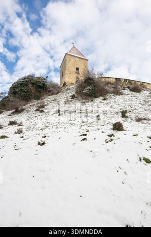 Une tour de pierre se trouve au sommet d'une colline enneigée, entourée d'arbustes clairsemés et d'un mur altéré. Le ciel bleu clair met en valeur un paisible fortre historique Banque D'Images