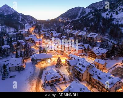 Vue aérienne des toits enneigés brillent chaleureusement contre les montagnes sombres de Vialattea, niché dans les Alpes italiennes, Claviere, Piémont, Italie. Banque D'Images