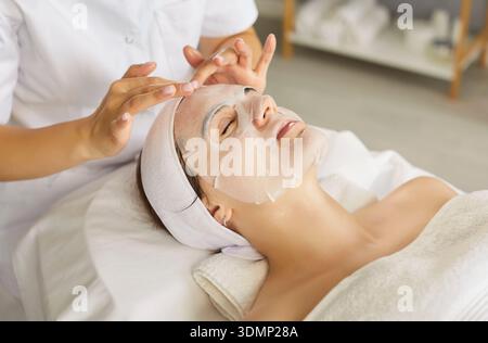 Esthéticienne ou cosmétologue appliquant un masque hydratant en feuille sur le visage de la jeune femme Banque D'Images