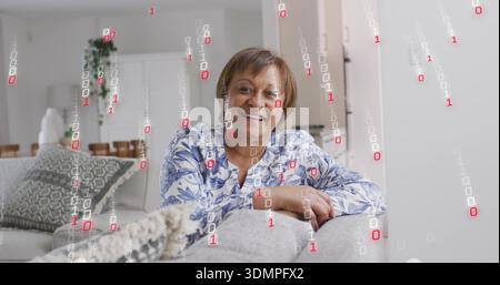 Femme mature souriante assise sur un canapé gris clair à la maison, portant un chemisier bleu avec superposition binaire Banque D'Images