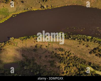 La vue aérienne montre une large rivière sombre courbant à travers la Lituanie rurale, avec des roseaux, des arbres dispersés, des prairies ensoleillées, des chemins boisés, et un petit étang à Banque D'Images