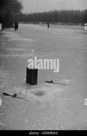 Slavyansk, RSS d'Ukraine, années 1960 Une scène hivernale tranquille de pêche sous glace sur une rivière gelée (probablement les Kazeny Torets). Au premier plan, de simples engins de pêche reposent sur la glace : deux petites cannes à pêche et une boîte à matériel rectangulaire qui sert également de siège. Un nouveau trou a été foré à proximité. En arrière-plan, des silhouettes d'autres pêcheurs se tiennent sur la vaste étendue enneigée, attendant une prise. La rive est bordée d'une forêt sombre dense. Cette photo d'archives atmosphérique capture la patience et la solitude des loisirs hivernaux dans la paisible région de Donetsk. Banque D'Images