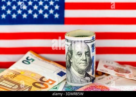 Gros plan macro d'un rouleau de billets en dollars américains au-dessus d'autres billets de banque en devises avec le drapeau des États-Unis d'Amérique en arrière-plan. Banque D'Images