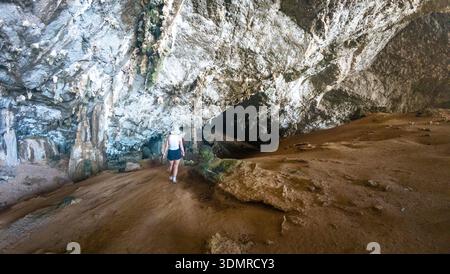 Randonneur marchant à l'intérieur d'une grande grotte calcaire avec des murs rocheux accidentés et un sol sablonneux, lumière naturelle à l'entrée de la grotte, voyage d'aventure et exploration c Banque D'Images