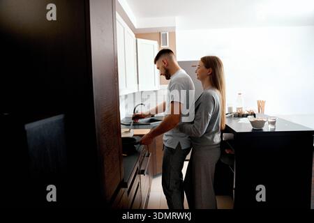 Un couple cuisine ensemble dans leur cuisine la nuit. L'homme est concentré sur une casserole tandis que la femme se tient proche et apprécie le moment. Banque D'Images