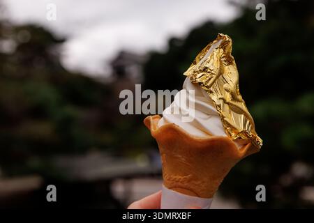 Un cône gaufrier contenant de la crème glacée blanche molle enveloppé dans une grande feuille de papier d'or comestible. L'arrière-plan présente un bokeh extérieur doux. Banque D'Images