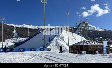 Livigno, Italie - 6 février 2026 : rampe de saut à ski olympique Milano Cortina 2026 avec ouvriers, projecteurs et Alpes enneigées dans la ville ensoleillée de Livigno, Italie Banque D'Images