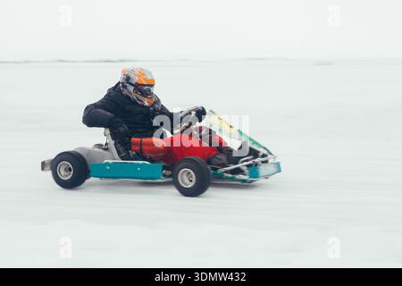 Coureur professionnel dans un casque et un équipement chaud conduisant un kart rapide sur une piste de rivière glacée blanche et enneigée lors d'une compétition de sport automobile hivernale intense pendant p Banque D'Images