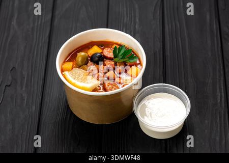 Soupe Solyanka avec saucisse, olives et citron dans un bol kraft avec de la crème sure sur une table sombre Banque D'Images