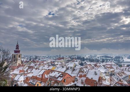 Scène de vieille ville enneigée avec des maisons aux toits rouges, une grande tour d'église et un ciel sombre et couvert. Une vue hivernale tranquille mettant en valeur l'architecture historique Banque D'Images