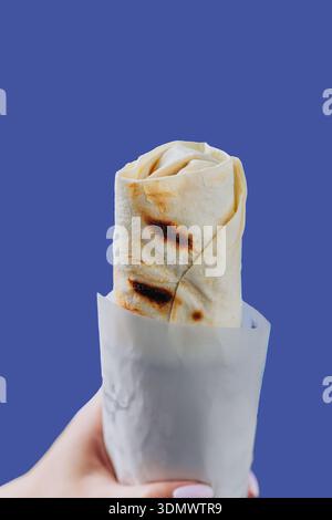 Main tenant shawarma enveloppé dans une pochette en papier isolé sur fond bleu Banque D'Images