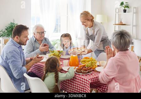 Grande famille multi-génération se réunissant autour de la table, applaudissant comme mère apporte une pizza fraîchement préparée. Banque D'Images