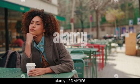 Une femme élégante aux cheveux bouclés est assise à une table de café verte en plein air, reposant son menton sur sa main avec un café à emporter à proximité. Ambiance urbaine calme Banque D'Images
