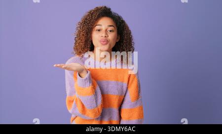 Portrait d'une jeune femme aux cheveux bouclés soufflant un baiser, portant un pull en tricot rayé lavande et orange sur un fond violet. Chaleureux, ludique, Banque D'Images