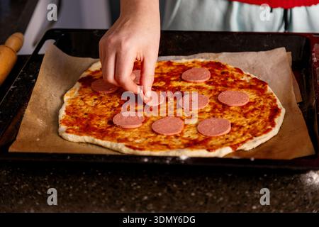 Une personne prépare une pizza maison en ajoutant des tranches de saucisse sur le dessus de la pâte, qui est recouverte de sauce tomate et de fromage, soulignant th Banque D'Images