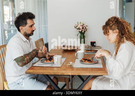 Un couple profite d'un petit déjeuner matinal confortable à une table en bois, avec de délicieuses pâtisseries et des tasses de café, avec un vase de fleurs ajoutant une touche de Banque D'Images