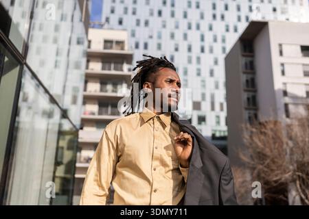 Homme d'affaires afro-américain avec des dreadlocks se promène en toute confiance dans un paysage urbain, tenant sa veste et regardant vers l'avant, entouré d'archite moderne Banque D'Images