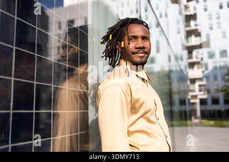 Homme d'affaires afro-américain avec des dreadlocks se tient confiant contre un mur de bâtiment réfléchissant dans un cadre urbain, portant une chemise beige. Il exsude Banque D'Images
