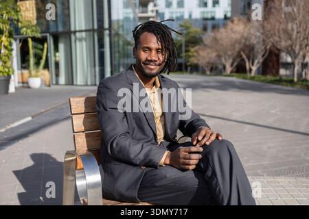 Homme d'affaires afro-américain avec des dreadlocks noirs assis en toute confiance sur un banc dans un cadre urbain, portant un costume et souriant chaleureusement à la caméra Banque D'Images