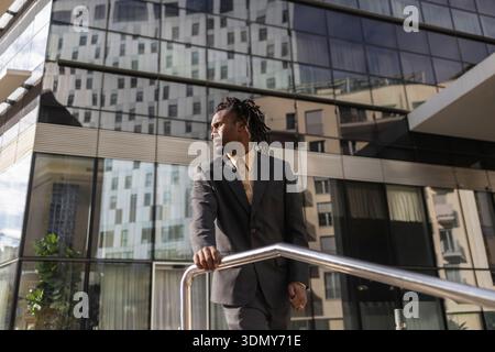 Homme d'affaires afro-américain avec des dreadlocks debout en toute confiance devant l'architecture en verre moderne, portant un costume formel, regardant soigneusement je Banque D'Images