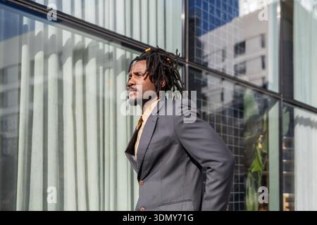 Homme d'affaires afro-américain avec dreadlocks se tient avec confiance devant un bâtiment en verre moderne, affichant professionnalisme et détermination dans un Banque D'Images
