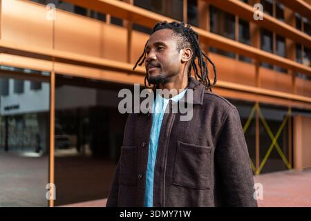 Homme d'affaires afro-américain avec des dreadlocks se tient en toute confiance dans un cadre urbain moderne, portant un manteau élégant, reflétant le professionnalisme et la suite Banque D'Images