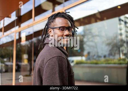 Homme d'affaires afro-américain avec des dreadlocks et des lunettes, regardant en arrière avec confiance tout en se tenant près d'un bâtiment vitré, capturant un sentiment de moder Banque D'Images