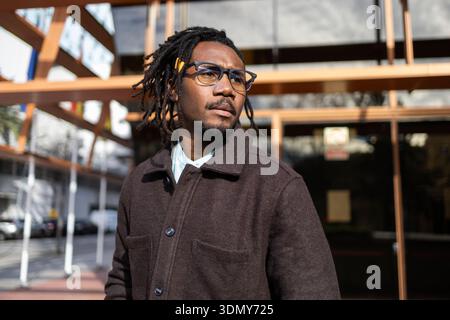 Homme d'affaires afro-américain avec des dreadlocks portant des lunettes et un manteau foncé, debout en toute confiance à l'extérieur devant un immeuble de bureaux en verre moderne Banque D'Images